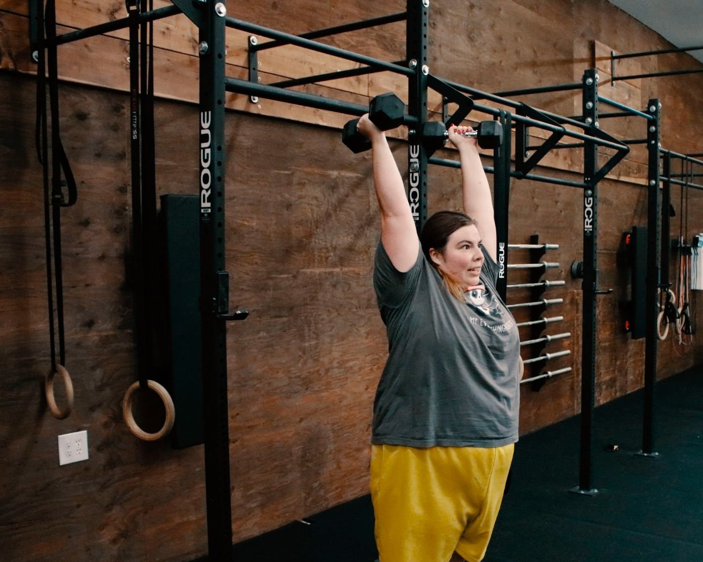 Woman lifting a dumbbell overhead in a gym with black metal frames and wooden wall