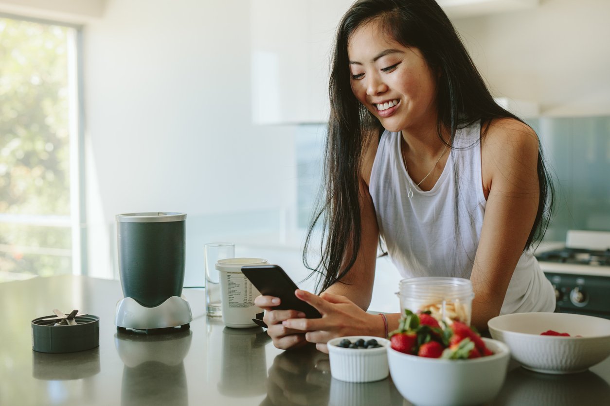 A woman smiling at her phone in a bright kitchen with bowls of fruit and a blender on the counter